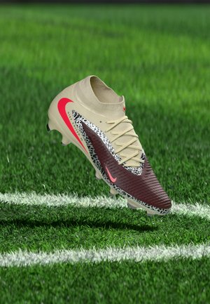 Beige og brune Nike-fotballsko med rød Swoosh og dyreprintdetaljer, svevende over grønt gress nær hvite banemerker.