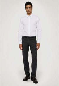 Bílá košile s dlouhým rukávem a límečkem na knoflíky, spárovaná s tmavě šedými kalhotami a černými botami. Jednoduchý, dobře střižený design s hladkou texturou.