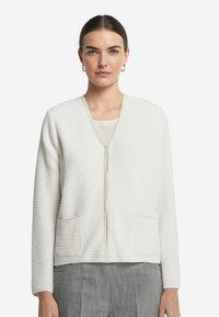 Weißer strukturierter Cardigan mit V-Ausschnitt, Vorderreißverschluss und zwei Taschen vorne. Verfügt über ein dezentes Muster und eine passende Borte.