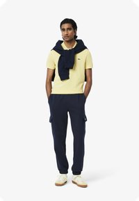 Geel polo-shirt, marineblauwe trui over de schouders, marineblauwe cargobroek en witte sneakers met beige accenten. Casual outfit.