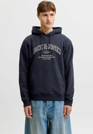 Młody mężczyzna w ciemnogranatowej bluzie z kapturem Jack & Jones i jasnoniebieskich jeansach, stojący na jednolitym szarym tle.