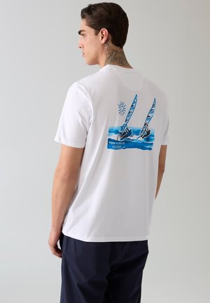 Mężczyzna z tatuażem na szyi, ubraną białą koszulkę z niebieską grafiką dwóch windsurferów i słońca, w ciemnych spodniach, zwrócony bokiem.