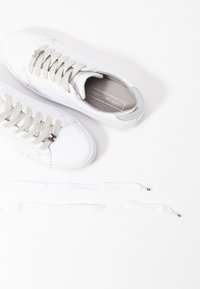 Pair of white Tommy Hilfiger sneakers with beige laces, branded metal eyelets, and an extra pair of white shoelaces on a white background.