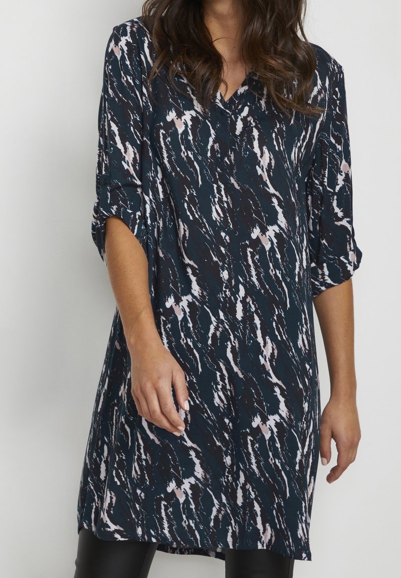 Femme portant une robe longue jusqu'aux genoux à manches longues avec un motif abstrait noir, blanc et rose clair, debout devant un fond gris uni.