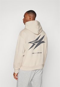 Felpa beige con un grande design grafico nero sul retro, maniche e orlo a costine. Abbinata a pantaloni della tuta grigi.