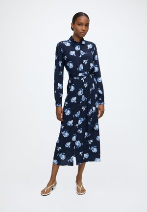 Femme portant une longue robe fleurie bleu marine avec des manches longues et une ceinture à la taille, debout devant un fond blanc uni.