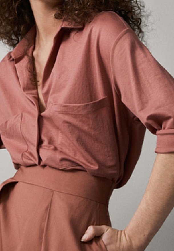 Femme portant une chemise ample rose poudrée boutonnée, rentrée dans un pantalon taille haute assorti, une main sur la hanche.