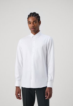 Langærmet hvid skjorte med knaplukning foran, spidse kraver og glat tekstur. Modellen har den på med mørke bukser.