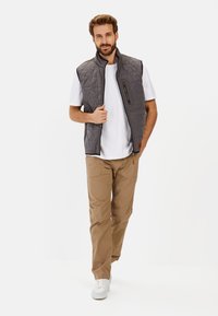 Grå quilted vest med høj krave, lynlåslommer, båret over en hvid t-shirt og khaki bukser. Inkluderer en læder accentetiket. Hvide sneakers.