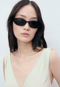 Black cat-eye sunglasses with a thin frame, worn by a model. The model has long dark hair and is dressed in a light green top.
