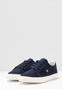 Námornické plátěné tenisky s bílými gumovými podešvemi a tkaničkami; mají malou zlatou značku na boku. Klasický design s nízkým střihem.