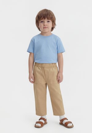 Ung barn med brunt hår står, iført en lyseblå t-shirt, beige bukser med elastik i taljen, hvide sokker og brune sandaler.