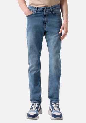 Mand iført lyseblå slim-fit jeans med hænderne i lommerne og blå og hvide sneakers, stående mod en ensfarvet baggrund.