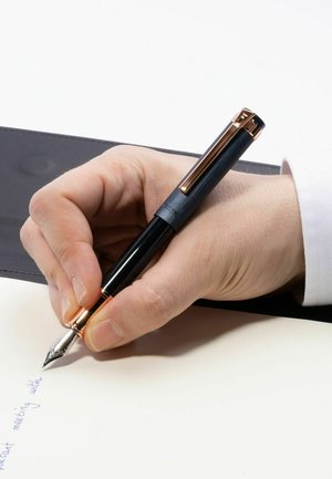 Fountain pen with a black and gold design held by a hand, writing on light paper. The pen has a glossy finish and ornate nib detail.