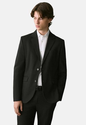 Blazer negro con un corte entallado, dos botones, solapas de muesca y un bolsillo en el pecho. Llevado sobre una camisa blanca de cuello y pantalones negros.