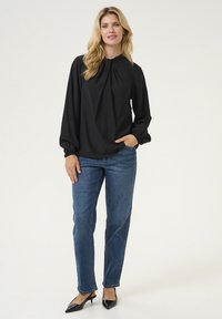 Blusa nera a maniche lunghe con collo a nodo, con maniche a palloncino. Abbinata a jeans in denim blu a vita alta e scarpe col tacco nere.