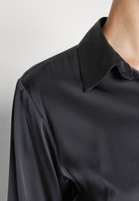 Zwarte satijnen blouse met een klassieke kraag, een soepele textuur en een getailleerde pasvorm. De stof heeft een lichte glans, die de details van het kledingstuk accentueert.
