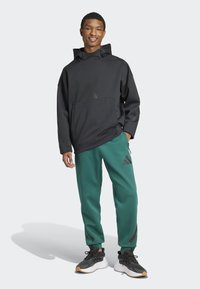 Μαύρη φούτερ με κουκούλα και μπροστινή τσέπη, διακοσμημένη με γεωμετρικά στοιχεία, συνδυασμένη με πράσινες φόρμες που διαθέτουν λογότυπο και πλαϊνά φερμουάρ, φορεμένη με μαύρα αθλητικά παπούτσια.