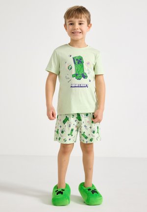 Fiú mosolyog világoszöld, Minecraft témájú pizsamában, Creeper grafikával és hozzáillő zöld Creeper papucsban, fehér háttér előtt áll.