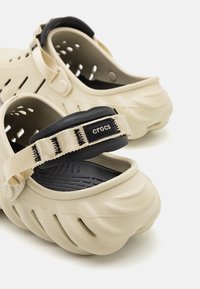 Des Crocs beiges avec des sangles de talon rembourrées noires, des trous de ventilation et des semelles texturées visibles en vue rapprochée de côté.