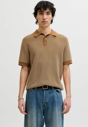 Mladi muškarac nosi smeđu polo majicu s uzorkom kratkih rukava i široke plave traperice na jednostavnoj svijetloj pozadini.