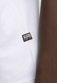 Camiseta de algodón blanca con una etiqueta negra de G-Star Originals. La imagen en primer plano muestra una textura suave y un diseño sencillo, sin patrones adicionales.