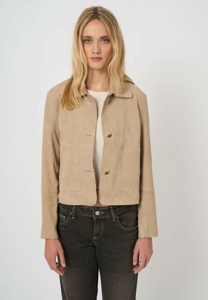 Vrouw met lang blond haar, gekleed in een beige jasje met knopen over een wit shirt en donkere spijkerbroek, staand tegen een effen lichte achtergrond.
