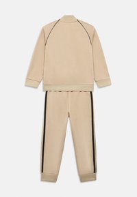 Bež trenirka od mekog tkanina, s jaknom na patentni zatvarač s crnim detaljima i odgovarajućim hlačama sa bočnim prugama i rebrastim manžetama.