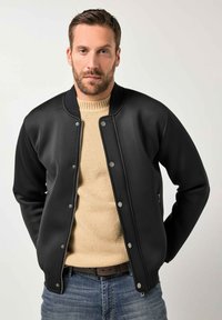 Giacca bomber nera con texture liscia, bottoni a pressione e colletto a coste, indossata sopra un maglione color cognac, abbinata a jeans blu.
