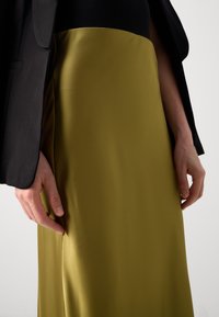 A close-up of an olive green satin skirt, with a smooth texture, paired with a black blazer. A hand is gently holding the fabric.