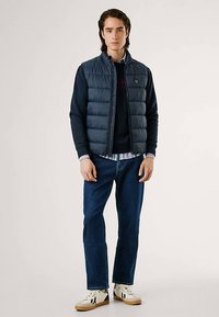 Colete acolchoado azul-marinho com fecho de correr à frente, por cima de uma camisola escura. Calças de ganga azul com corte reto e ténis brancos com detalhes pretos. Design minimalista.