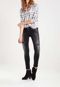 Chemise à carreaux gris et blanc avec manches retroussées, associée à un jean skinny noir déchiré aux genoux, et des bottines noires à talon large.
