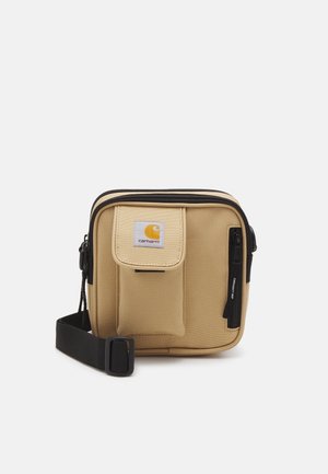 Kleine beige Carhartt schoudertas met voorvak, zwarte ritssluiting en verstelbare zwarte riem op witte achtergrond.