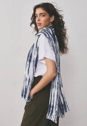 Young woman with curly hair wears white shirt and dark pants, draped with a long white scarf with blue dotted pattern.