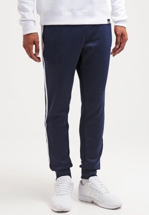 Person wearing navy blue jogger pants with white side stripes, white sneakers, and a white sweatshirt, standing on a white background.