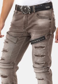 Abgenutzte braune Jeans in schmaler Passform mit mehreren Rissen, gestickten Details und einem CIPO & BAXX Logo-Patch. Begleitet von einem strukturierten schwarzen Gürtel.