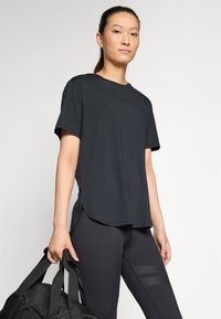 Black short-sleeve t-shirt, soft fabric, regular fit, slight drop hem, paired with black leggings featuring textured accents. Carrying a duffel bag.