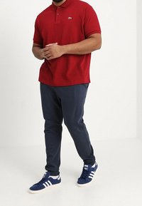 Polo rouge à manches courtes et col, arborant un petit logo. Assorti à un pantalon marine et des baskets bleues avec des rayures blanches.