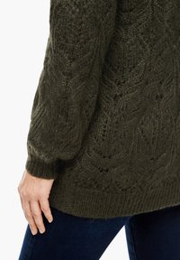 Dunkelgrüner, gestrickter Pullover mit komplizierten Zopfmuster, gerippten Bündchen und Saum; weicher, strukturierter Stoff. Hand ruht auf der Hüfte über einer Jeans.