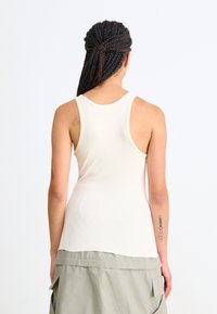 Krém színű tank top, versenyhátsó kialakítással, bordás anyagból készült. Lazább illeszkedéssel és ívelt alsó résszel rendelkezik, zöld, rétegelt szoknyával párosítva.