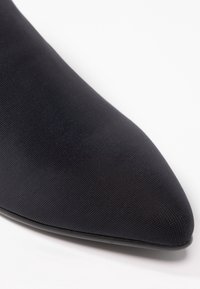 Gros plan sur un bout de chaussure en tissu noir pointu avec une surface finement texturée sur fond blanc.