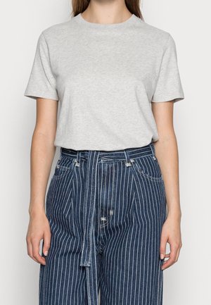 Kvinde iført en ensfarvet lysegrå kortærmet t-shirt, stukket ned i højtaljede blå jeans med striber og et matchende bælte.