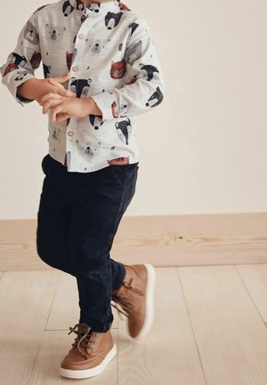 Barn iført skjorte med dyreprint, mørke bukser og brune snørestøvler, stående indendørs på lyst trægulv.