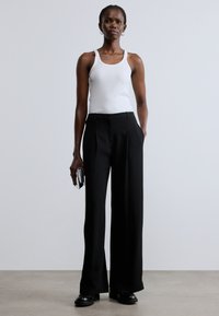 Fekete, széles szárú nadrág, szabott fazonú, sima textúrával és hajtásos elejjel. Fehér trikóval és elegáns cipővel párosítva.