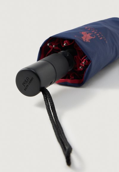 Guarda-chuva azul-marinho com uma pega preta e interior vermelho. Possui uma alça trançada e ferragens metálicas para durabilidade. Logotipo impresso na pega.