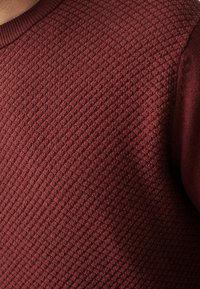 Pull texturé marron avec un col côtelé, un motif tissé et des accents en tissu uni sur les manches. Tissu doux avec un aspect structuré.