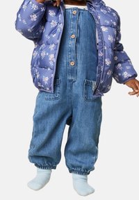 Maluch ubrany w jasnoniebieską kwiatową kurtkę puchową, dżinsowe ogrodniczki z guzikami i białe skarpetki, stoi na białym tle.