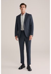 Completo sartoriale navy realizzato in tessuto testurizzato, abbinato a una camicia di colore chiaro. Il completo presenta due bottoni e pantaloni slim-fit, accompagnati da scarpe nere.