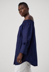 Camicia navy a spalle scoperte con scollatura arricciata, maniche lunghe, polsini con bottoni e spacchi laterali. Realizzata in tessuto leggero.