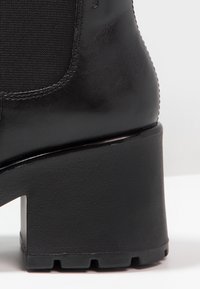 Bottine en cuir noir avec panneau latéral élastique et talon bloc épais, vue de côté mettant en avant le talon et les détails des coutures.